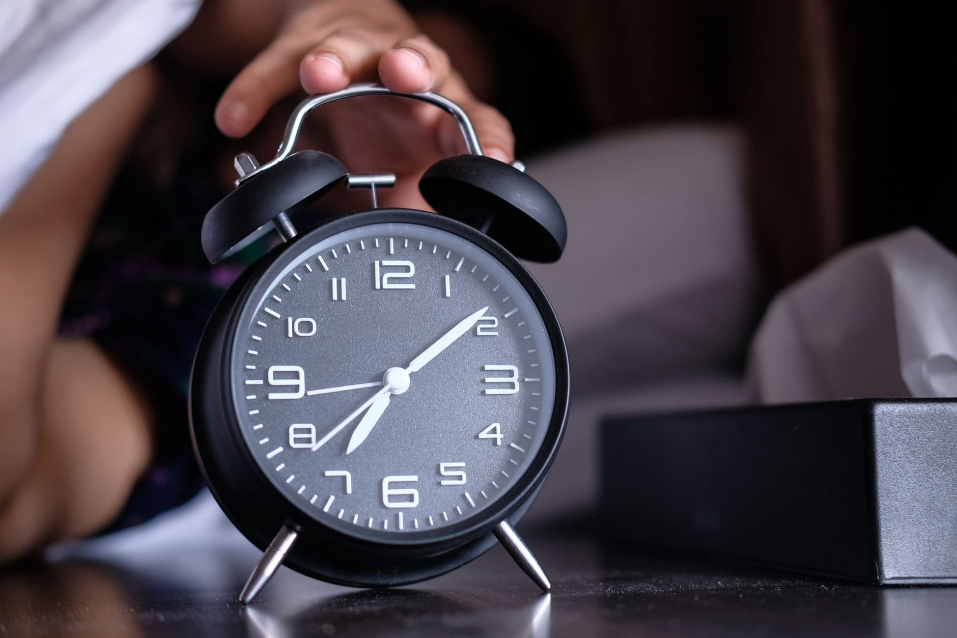 A hand reaching for an alarm clock on a bedside table